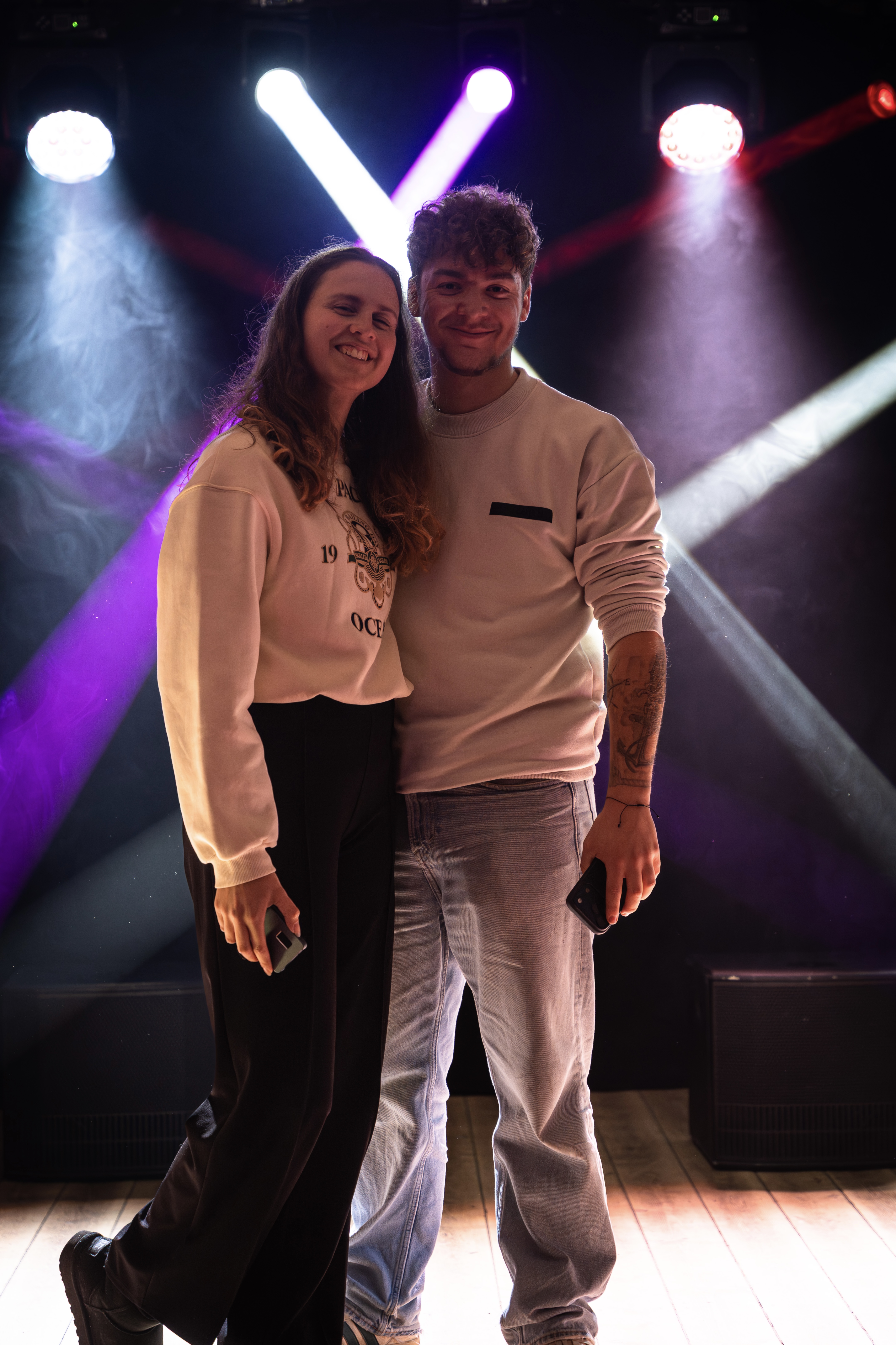 Jennifer und Christofer lächelnd auf einer Bühne im bunten Scheinwerferlicht